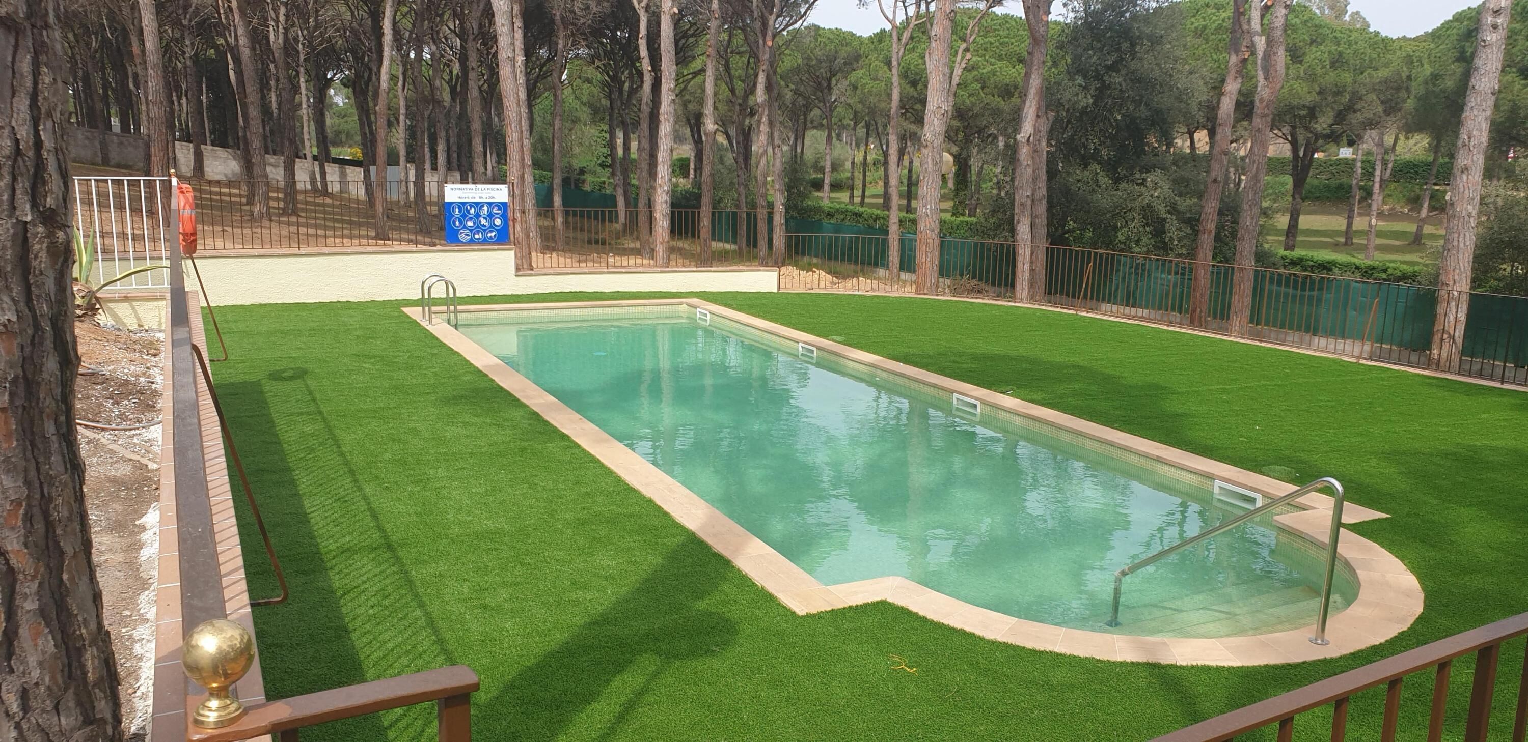 Una piscina rettangolare circondata da un'area di prato sintetico verde, una recinzione in metallo e alberi sullo sfondo.