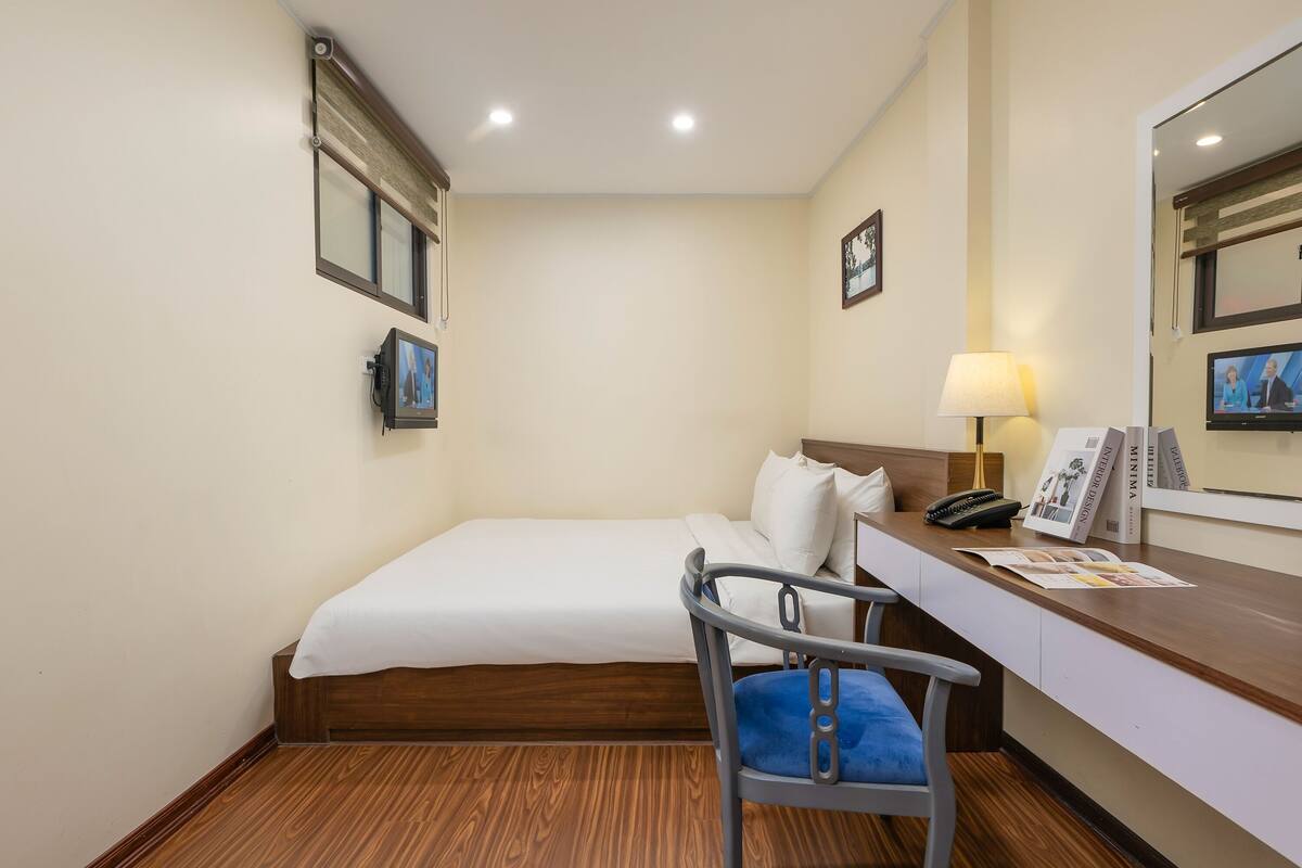 Une chambre d’hôtel avec un lit simple, un bureau avec une lampe, une chaise et un téléviseur fixé au mur.