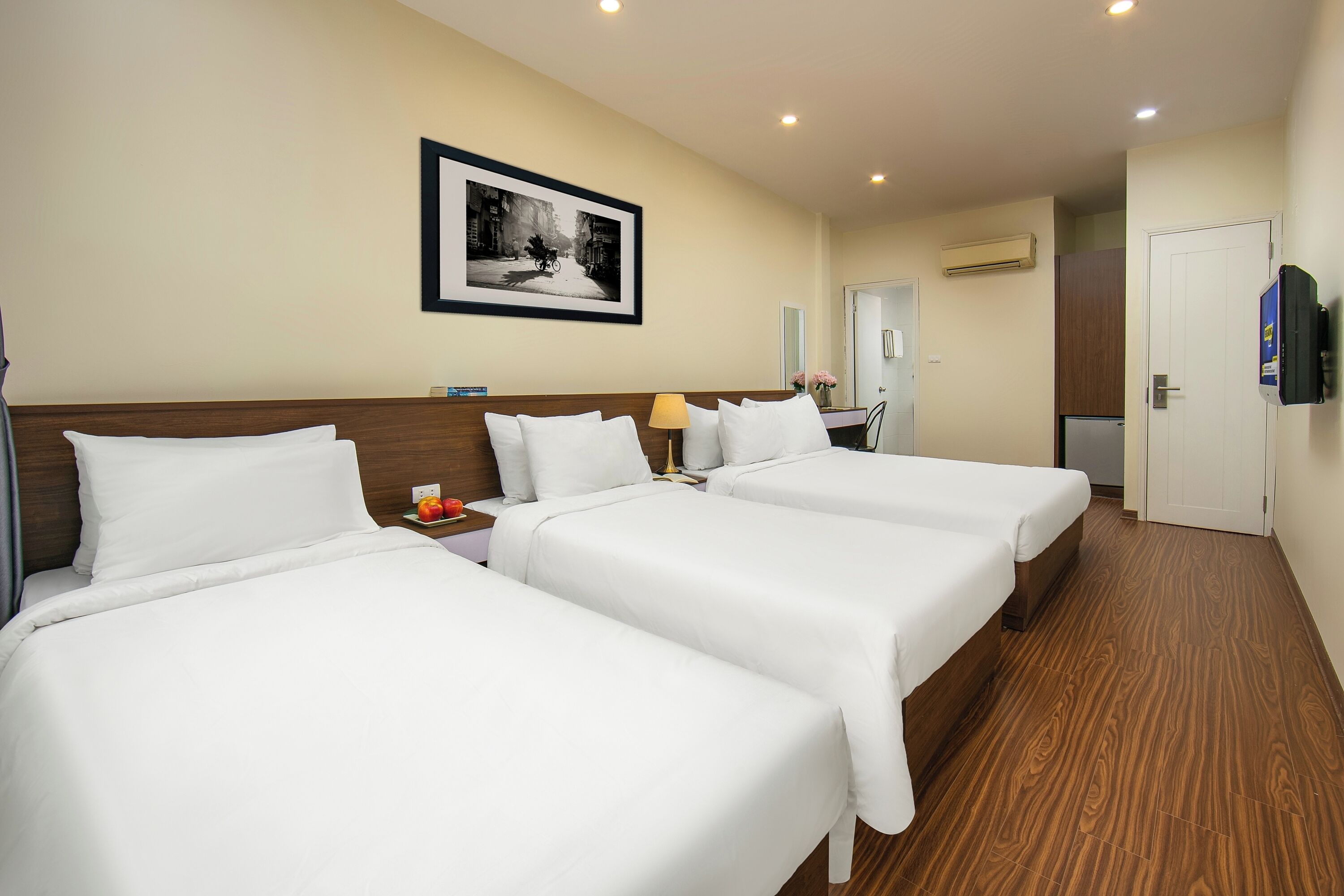 Habitación de hotel con tres camas individuales, cabecera de madera, un cuadro en la pared, unidad de aire acondicionado y un televisor montado.