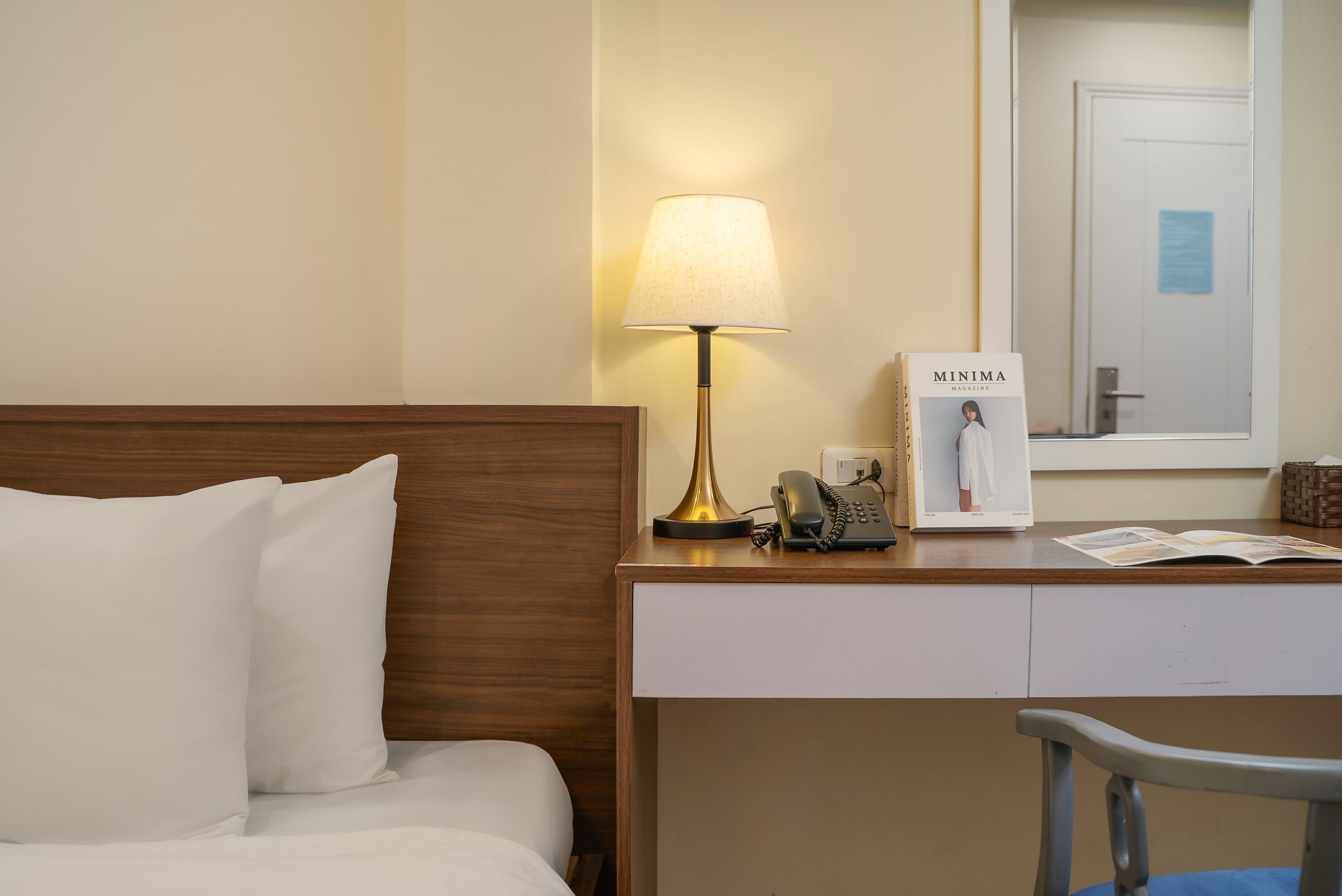 A hotel room with a bed, a desk with a lamp, a telephone, and a magazine.