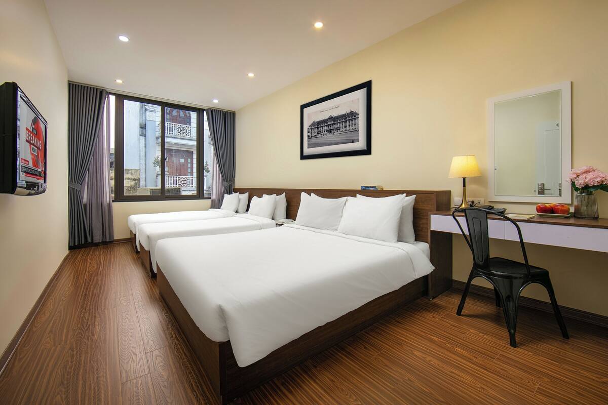Une chambre d’hôtel avec deux lits, un bureau, une chaise, une fenêtre avec des rideaux, un tableau encadré au mur et une lampe.