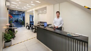 Een receptiebalie met een laptop, een telefoon en een tijdschrift. Er staan stoelen en tafels met planten op de achtergrond.