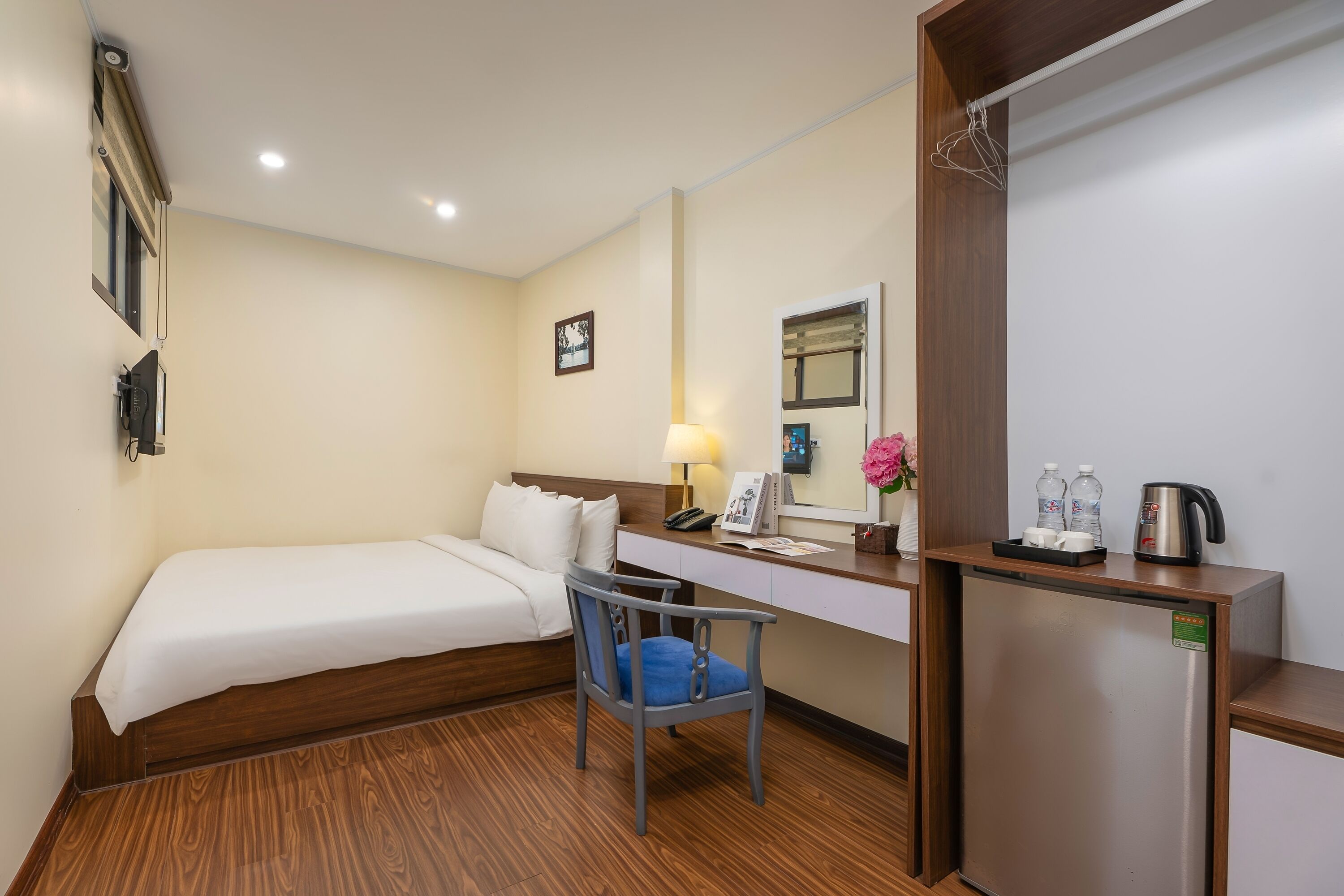 A hotel room with a bed, a desk with a lamp, a chair, a refrigerator, and a small shelf with items.