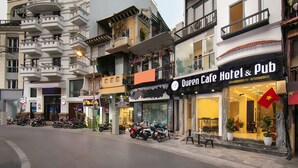 Straatbeeld met een hotel genaamd 'Queen Cafe Hotel & Pub', met een etalage met een groot glasraam en een rode vlagdecoratie.