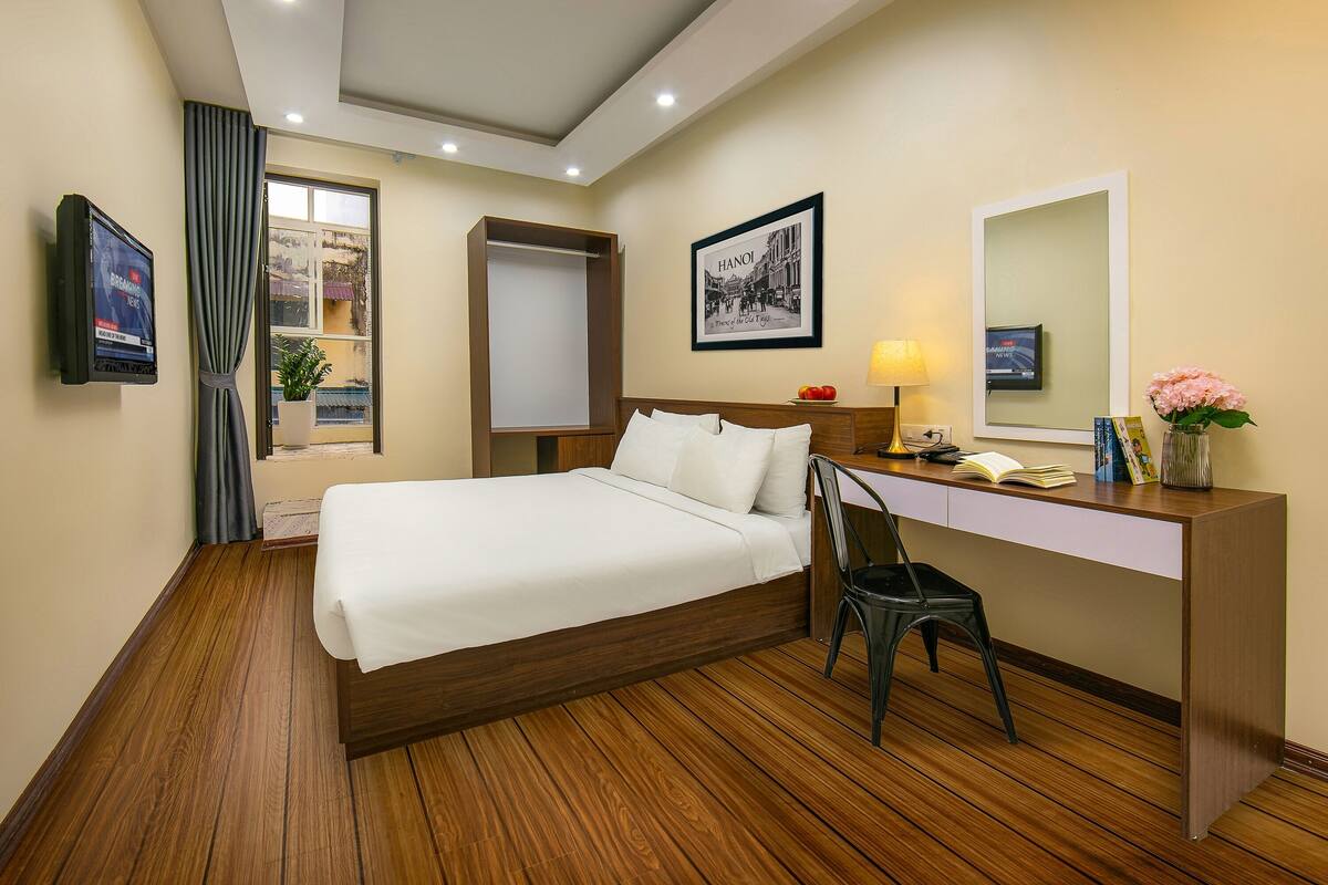Une chambre d’hôtel comprenant un lit, un bureau, une chaise, une télévision, une fenêtre avec des rideaux et un tableau encadré au mur.