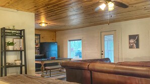 A cozy room with a wooden ceiling, a large leather couch, a bed with a blanket, a television, and a bookshelf.