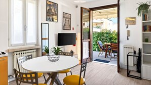 A room with a round dining table, chairs, a flat-screen TV, a bookshelf, and a door leading to an outdoor area with a table and chairs.