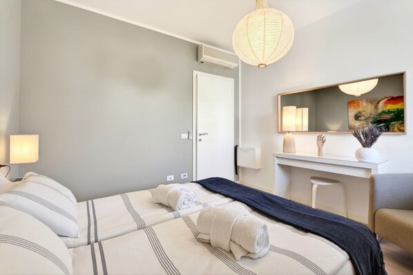 A hotel room with two beds, a desk, a chair, a mirror, and a hanging light fixture.