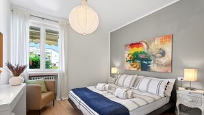 A bedroom with a bed, bedside tables, a desk, a chair, a window with curtains, and a painting on the wall.
