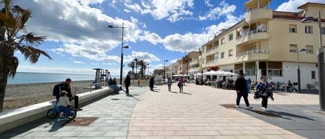 A paved promenade with a beach in the background, palm trees, and buildings with balconies.
