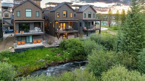 Une maison moderne à plusieurs étages, dotée d’un patio, d’un petit ruisseau et bordée par une forêt.