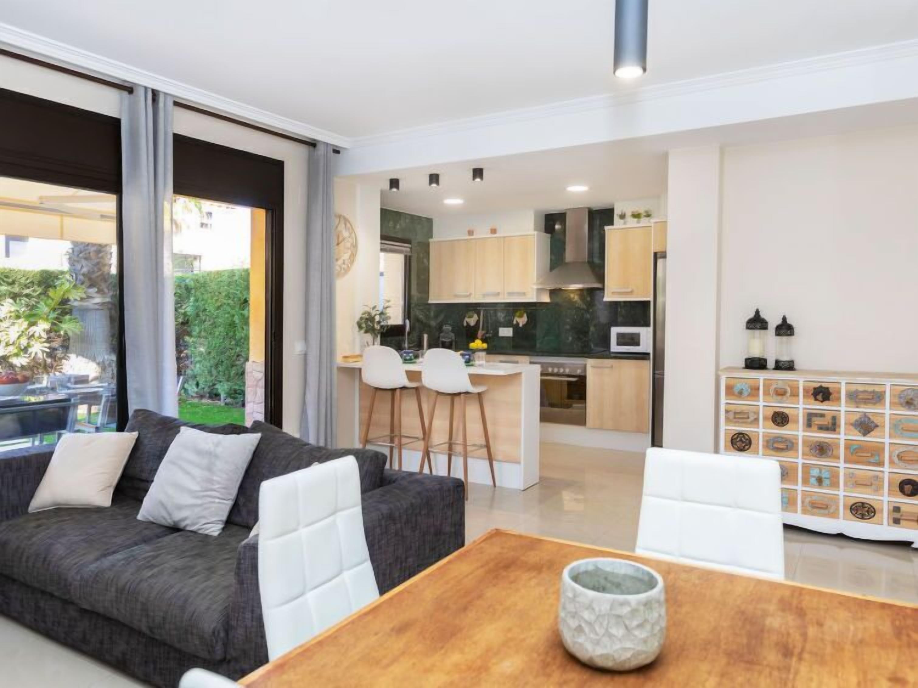A modern living room with a grey sofa, a wooden coffee table, and a view of a kitchen and dining area.