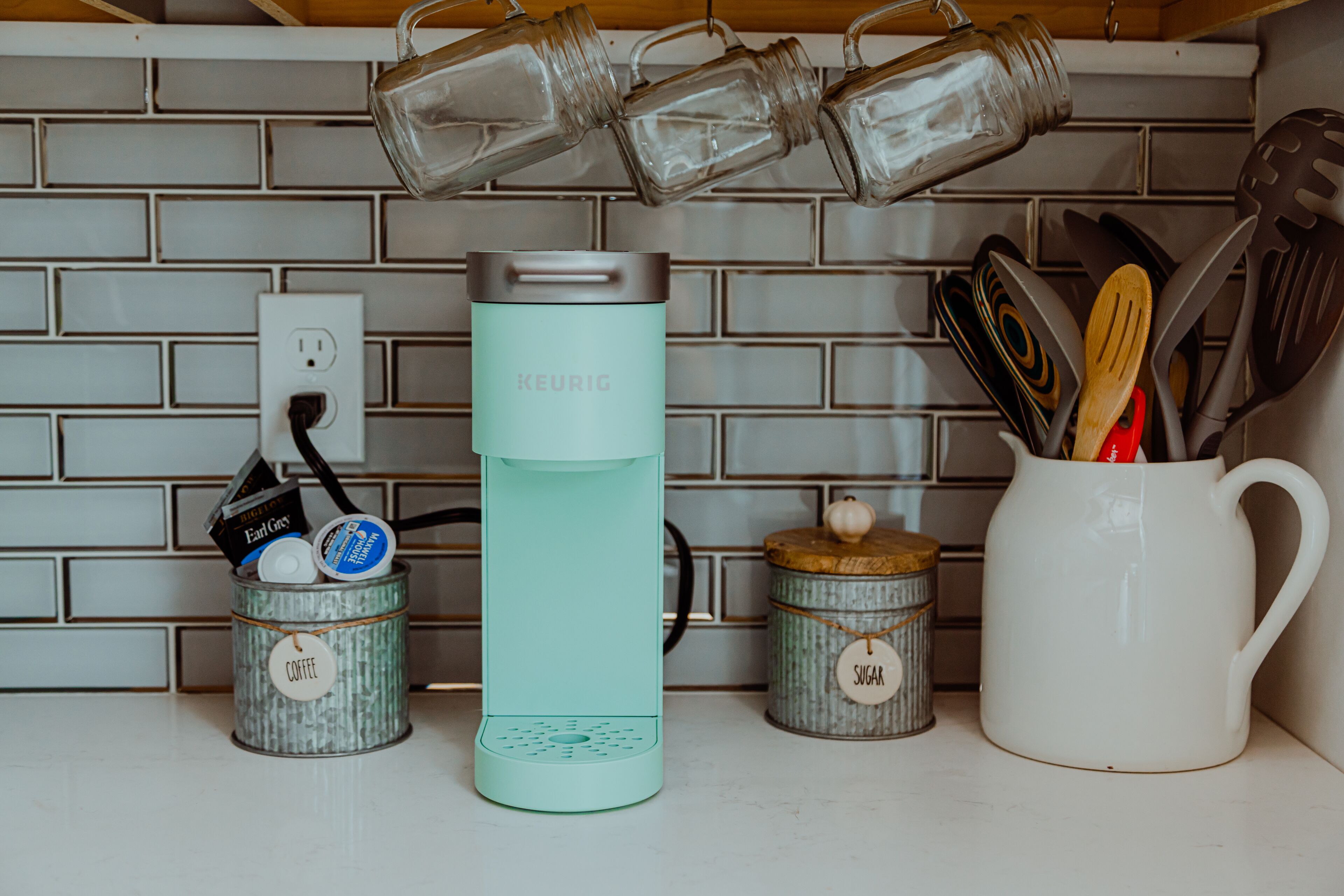 En køkkenbordplade med en teal Keurig kaffemaskine, to metalbeholdere mærket 'kaffe' og 'sukker', samt en hvid kop med bestik.