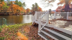 Une terrasse en bois, agrémentée de rampes blanches, surplombe un lac paisible, bordé d’arbres aux couleurs automnales.