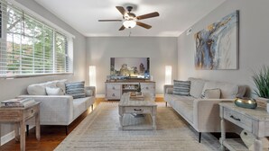 Un salon moderne doté d’un ventilateur de plafond, d’une grande fenêtre avec stores, d’un tableau représentant un paysage urbain et d’un design minimaliste.