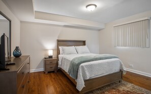 A bedroom with a wooden bed, a bedside table with a lamp, a dresser, and a large window with blinds. - Chicago charm! Quiet and inviting ! (Chicago)