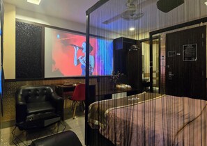 Une chambre d’hôtel avec un grand lit, un écran de télévision sur lequel on voit une personne, un fauteuil rouge et un fauteuil en cuir noir.