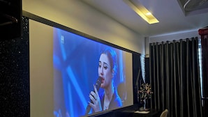 Habitación de hotel con un televisor de pantalla plana grande montado en la pared, una pequeña zona de estar con un banco y una silla, una mesita con un florero y una cama con cubrecolcha rojo y blanco.