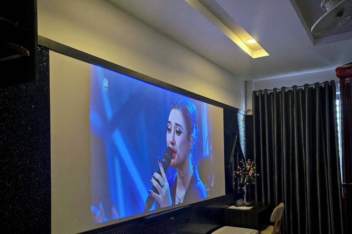 Habitación de hotel con un televisor de pantalla plana grande montado en la pared, una pequeña zona de estar con un banco y una silla, una mesita con un florero y una cama con cubrecolcha rojo y blanco.