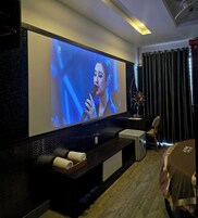 Habitación de hotel con un televisor de pantalla plana grande montado en la pared, una pequeña zona de estar con un banco y una silla, una mesita con un florero y una cama con cubrecolcha rojo y blanco.