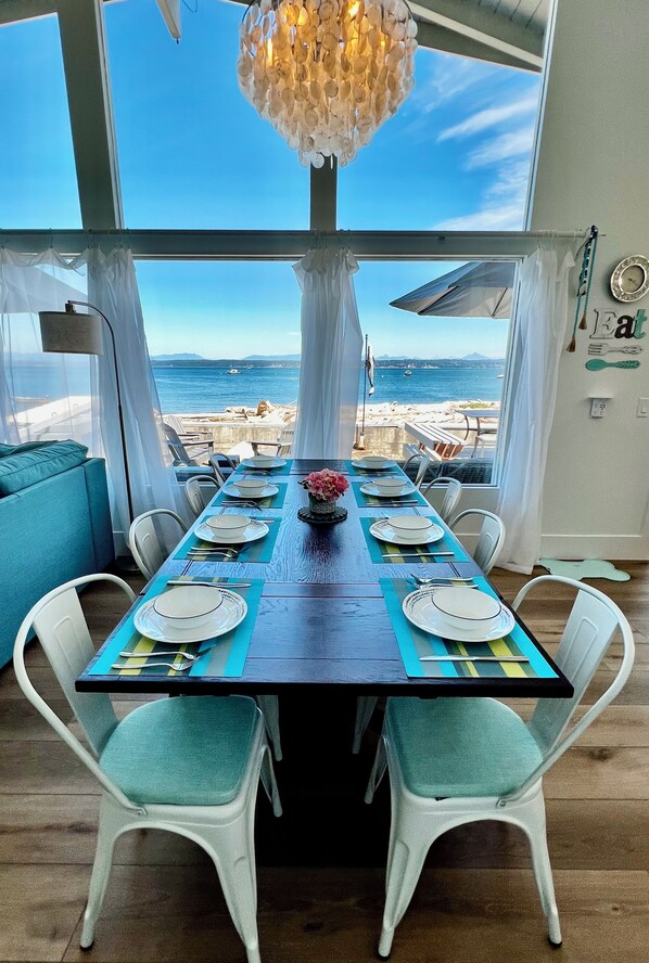 Zona pranzo con vista sul mare, tavolo in legno apparecchiato con piatti e posate, sedie bianche e un lampadario.