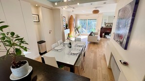 A modern dining area with a white table set for four, a chandelier, and a view of the outdoors through a large window. - Splendid House, Jacuzzi and Private Gardens Paris (Boulogne-Billancourt)