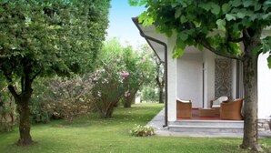 A covered outdoor seating area with a white wall and a patterned design, surrounded by lush greenery and trees.