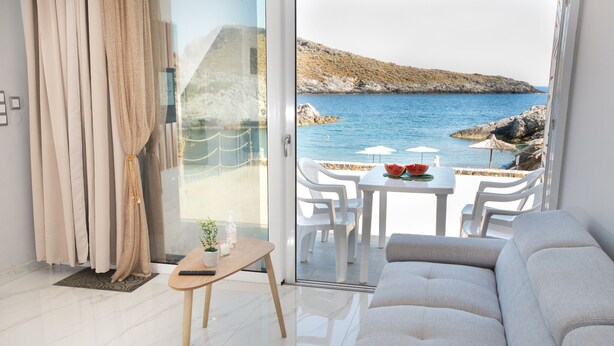 A modern living room with a large sofa, a small table with a plant, and a view of the sea through a glass door. - Beachfront Studio Kyma East (Faistos)