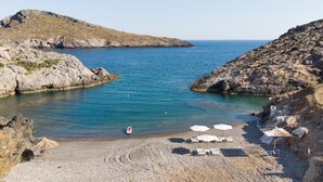 A rocky beach with clear blue water, a small boat, and beach umbrellas. - Beachfront Studio Kyma East (Faistos)