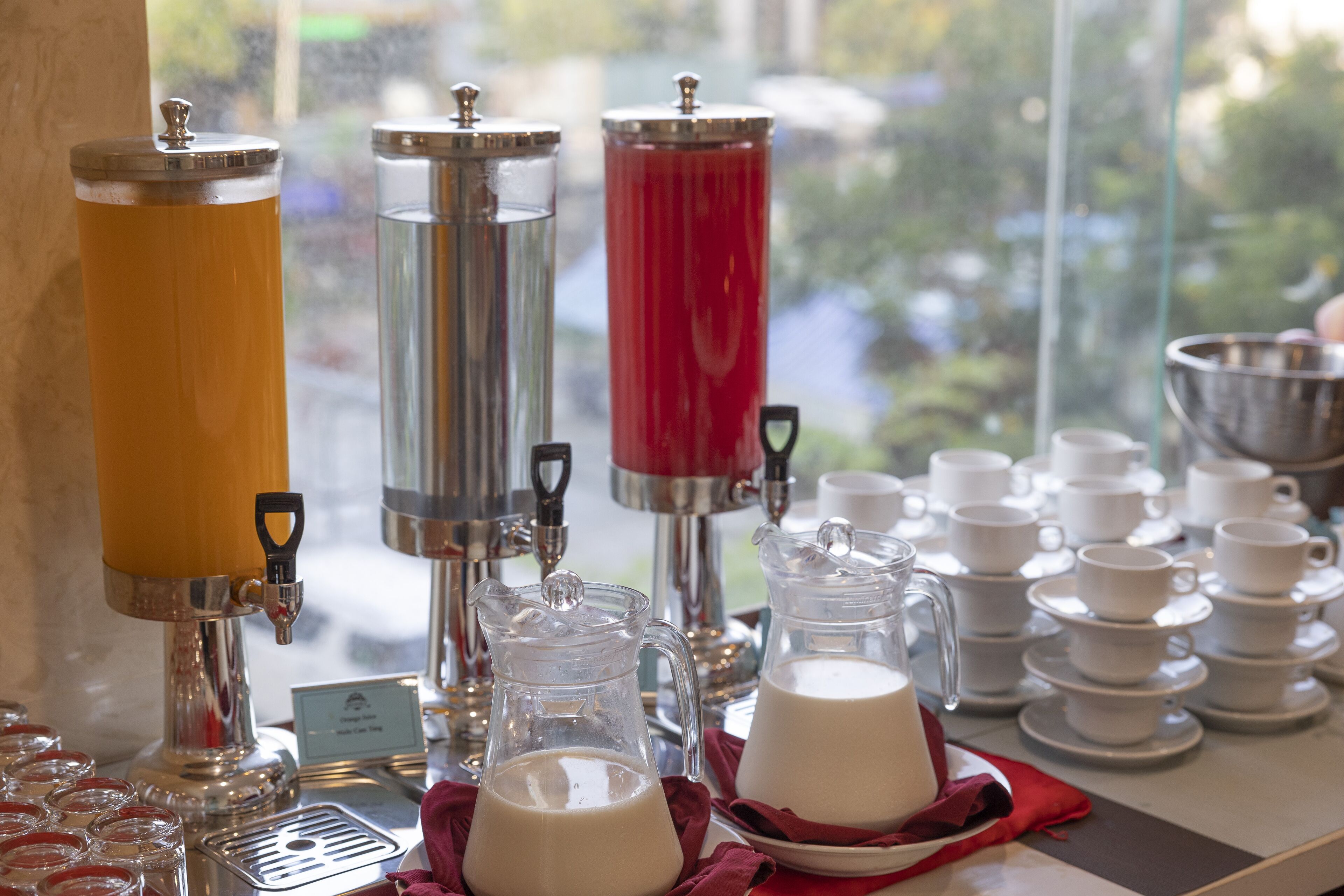 Een drankstation met schenkapparatuur en kannen, een stapel witte bekers, en uitzicht op een raam.