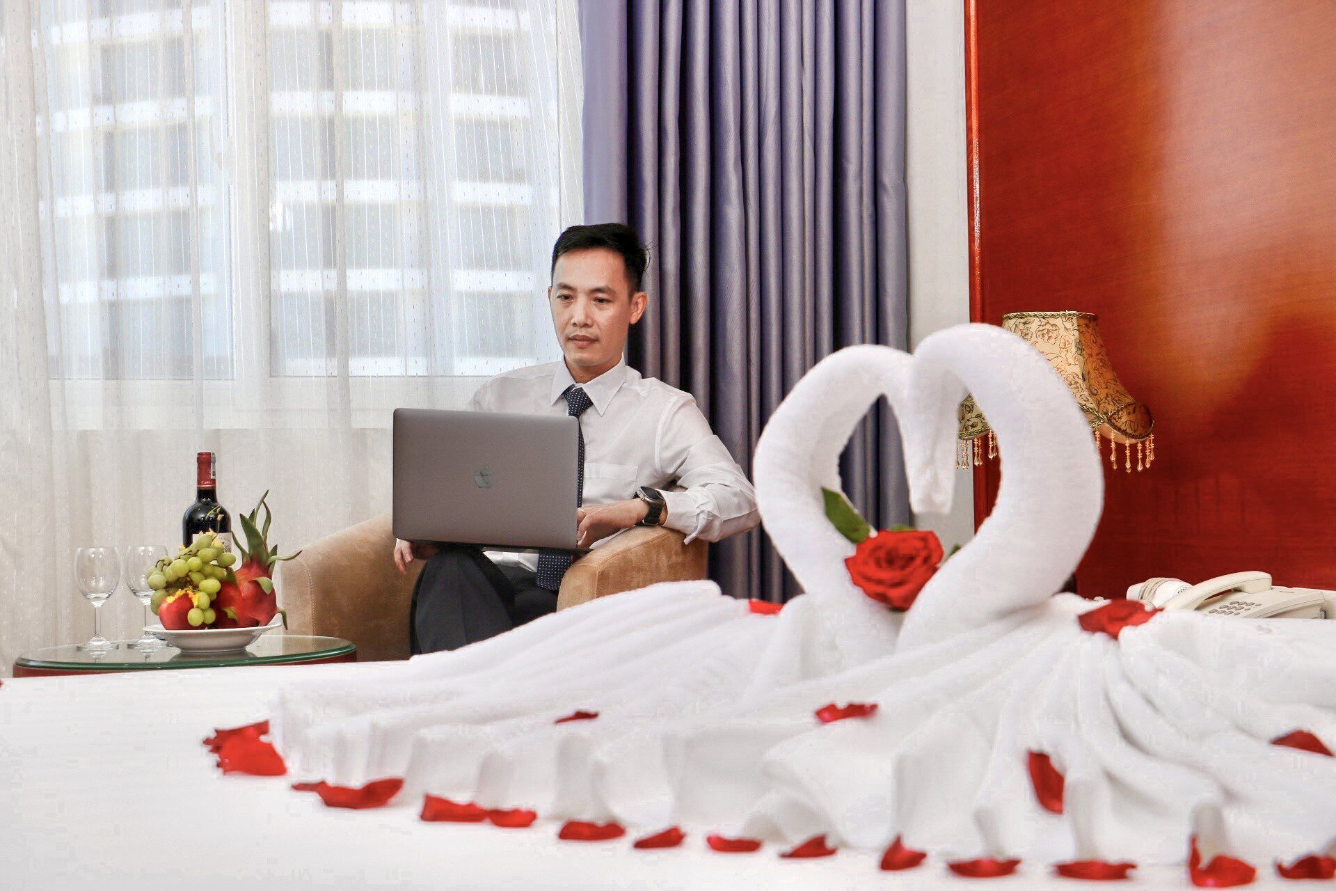 A man sitting on a bed with a laptop, a bouquet of flowers, and a swan-shaped decoration.