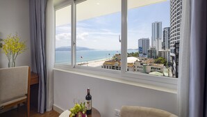 Una stanza con una grande finestra che si affaccia su una spiaggia e sullo skyline della città, un tavolino con una bottiglia e grappoli d'uva, e una sedia.
