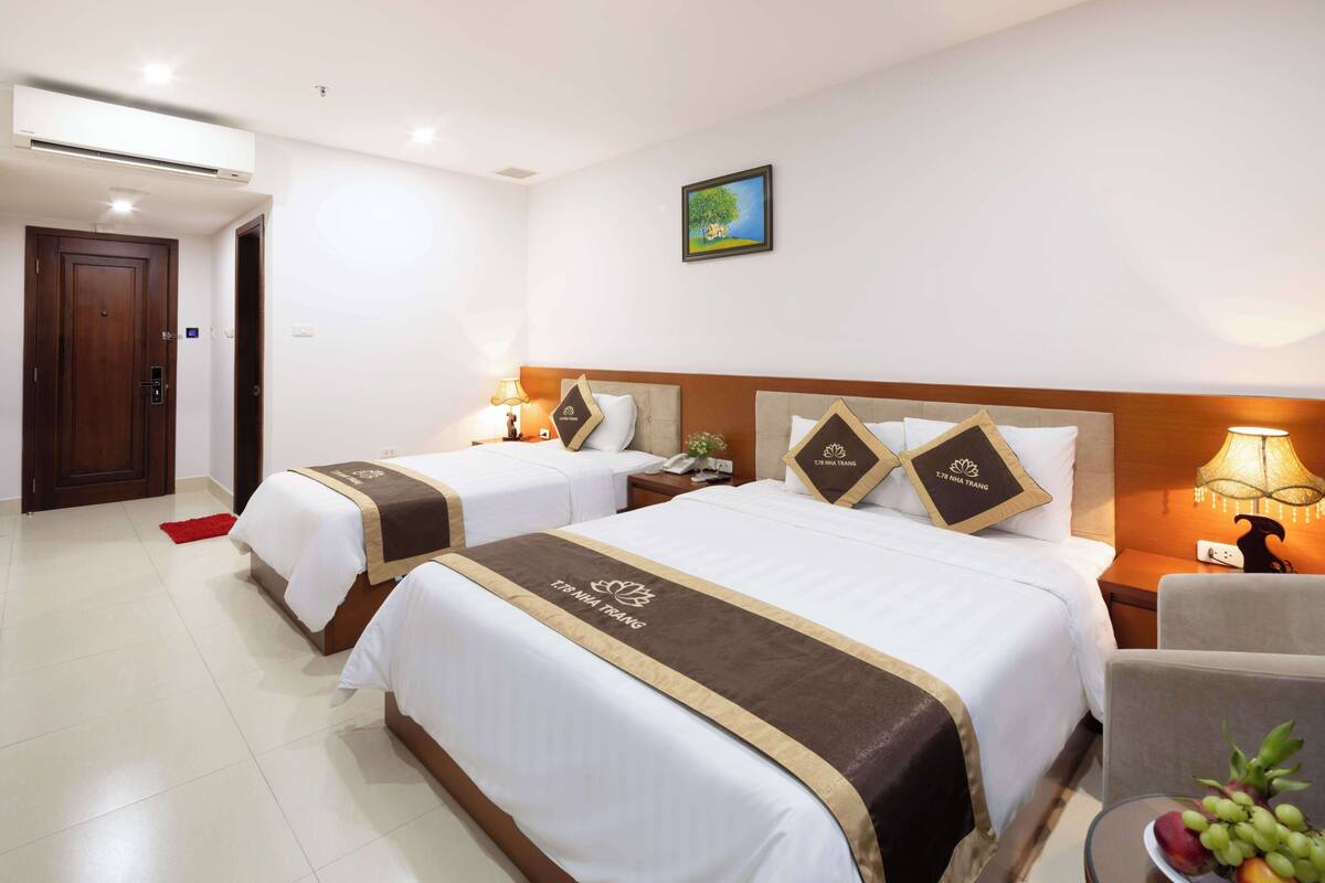 A hotel room with two beds, a wooden door, a nightstand with a lamp, a small table with fruit, and a painting on the wall.