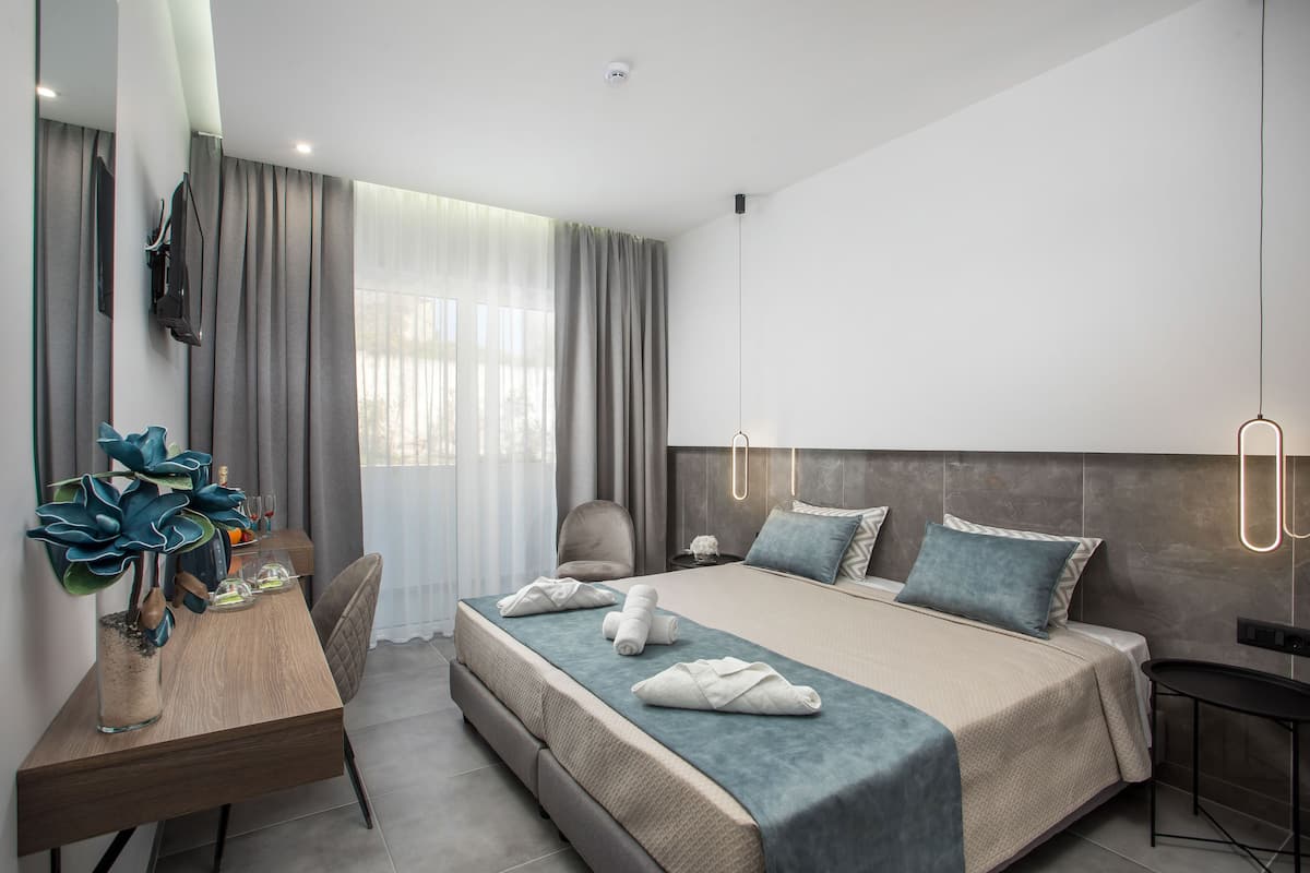 Une chambre d’hôtel moderne, dotée d’un grand lit, d’un bureau avec une chaise, d’une plante en pot et offrant une vue sur l’extérieur à travers de fines rideaux.