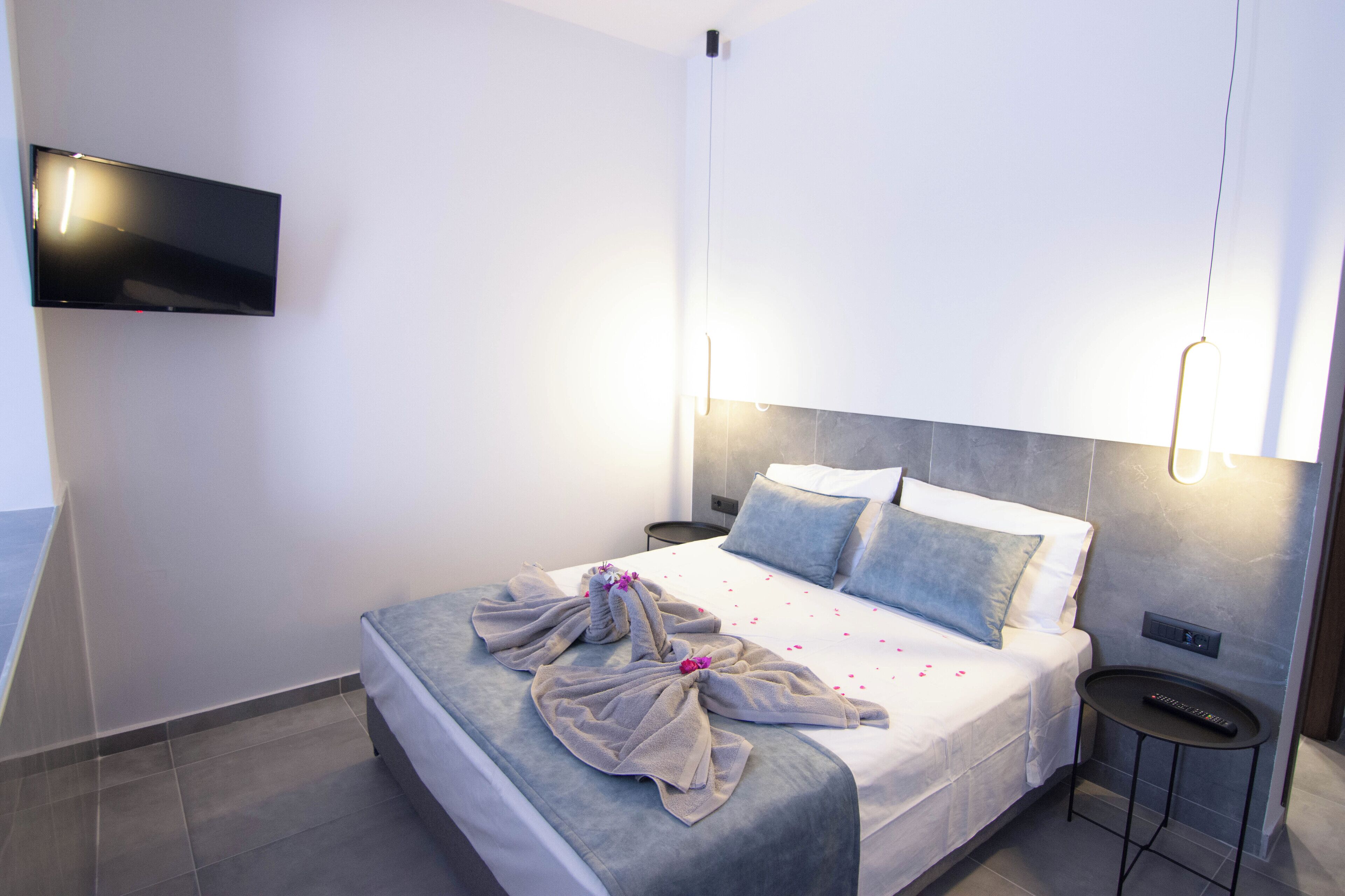 A modern hotel room with a bed, bedside tables, a flat-screen TV mounted on the wall, and a wall-mounted light fixture.