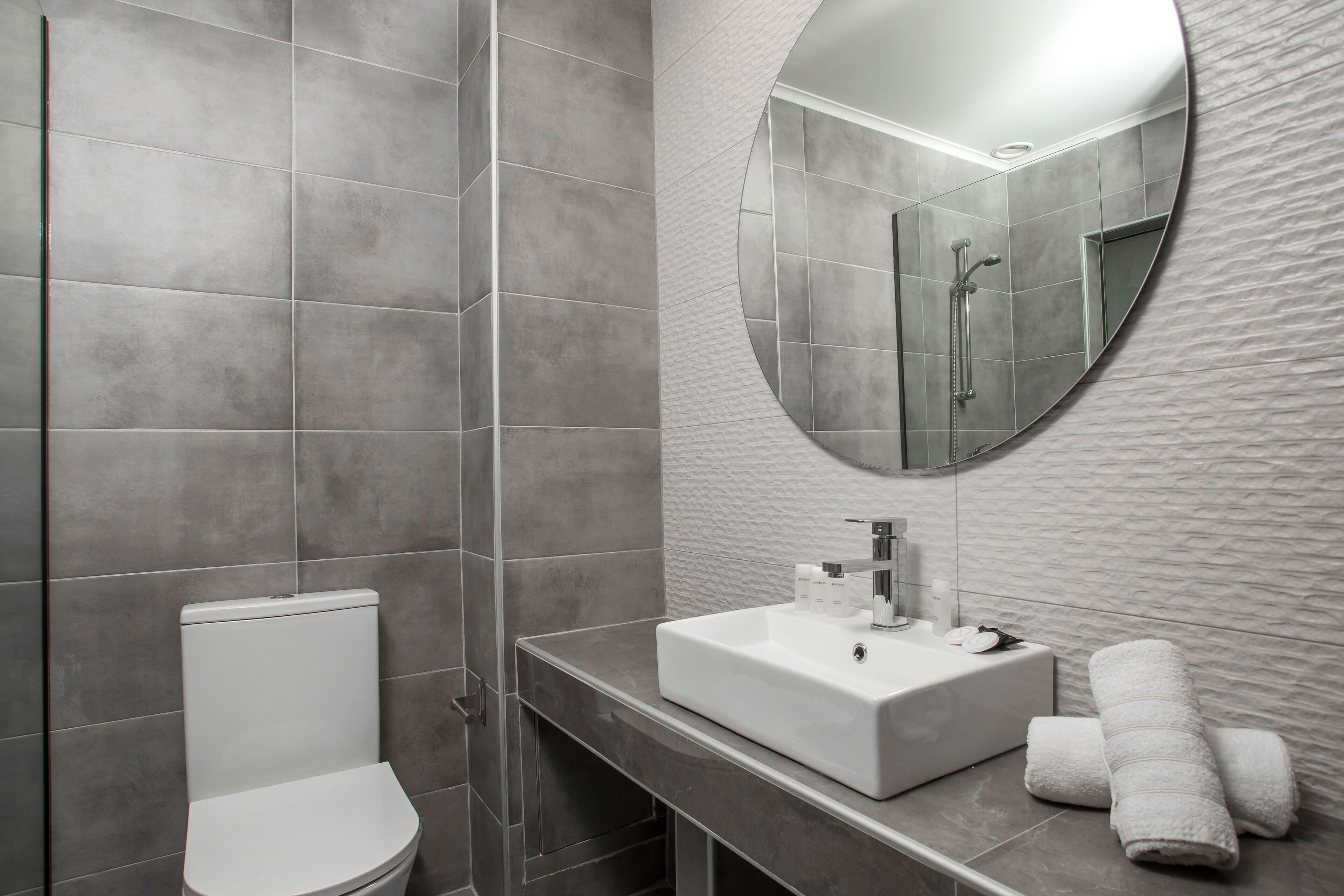 Un baño moderno con un inodoro blanco, un lavamanos rectangular y un espejo redondo.