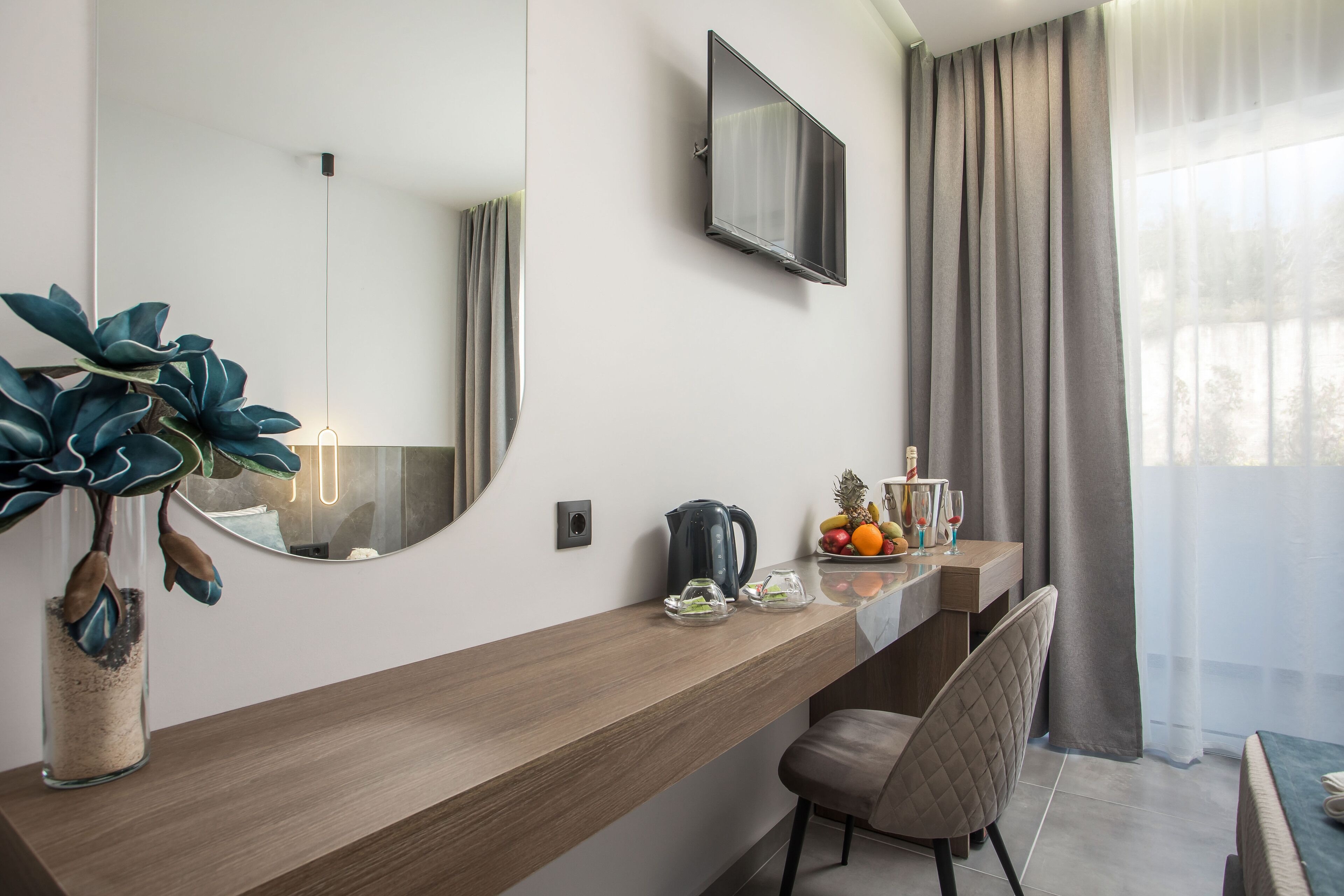 Ein modernes Hotelzimmer mit Schreibtisch aus Holz, einem großen Spiegel, einem Flachbildfernseher und Blick ins Freie durch leichte Vorhänge.