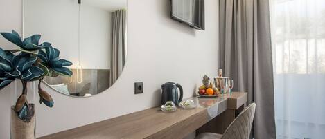 Ein modernes Hotelzimmer mit Schreibtisch aus Holz, einem großen Spiegel, einem Flachbildfernseher und Blick ins Freie durch leichte Vorhänge.