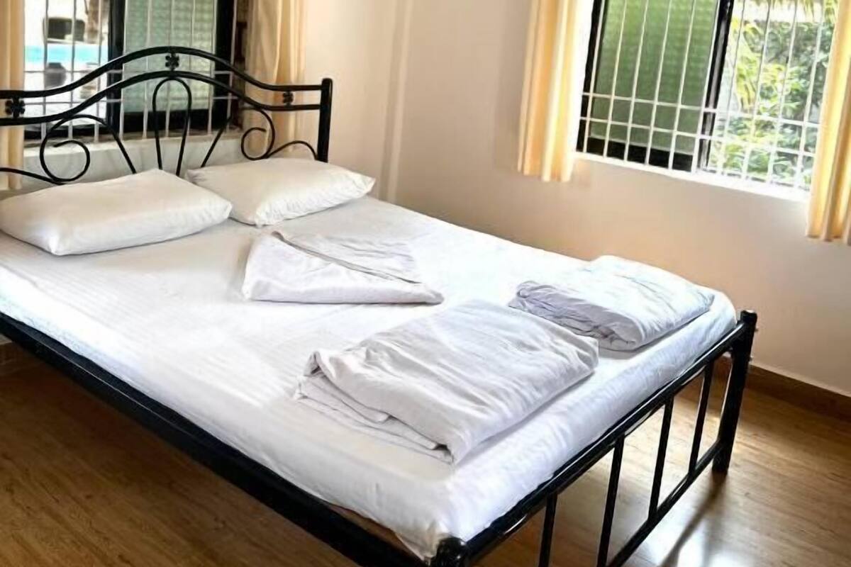 Ein Einzelbett mit Metallgestell, weißer Bettwäsche und zwei mit Vorhängen versehenen Fenstern.