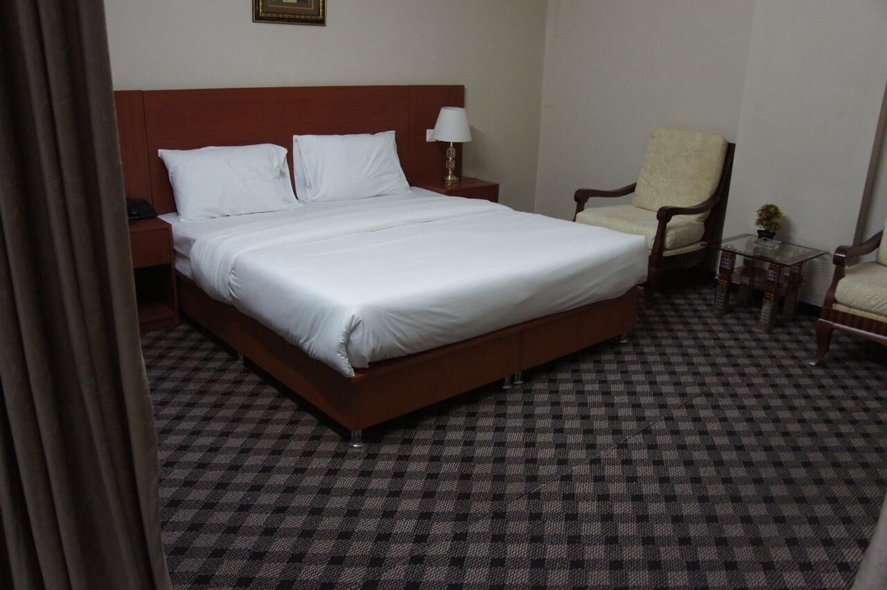 A hotel room with a large bed, a nightstand, a chair, and a small table with a lamp.