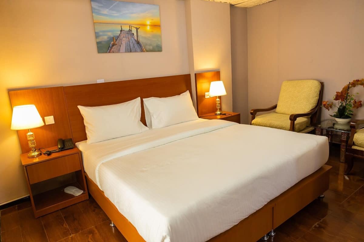 Hotellihuone, jossa on suuri sänky, kaksi yöpöytää valaisimineen, tuoli ja taulu seinällä.