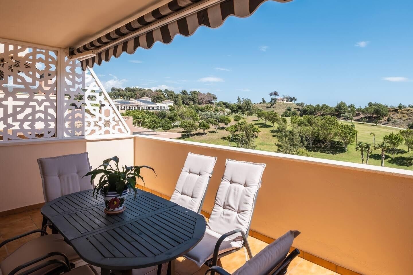 Un balcó amb una taula i dues cadires, una planta en test i vista cap a un camp de golf i edificis a la distància.
