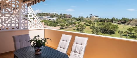 Un balcón con una mesa, dos sillas, una planta en maceta y vistas a un campo de golf y edificios a lo lejos.