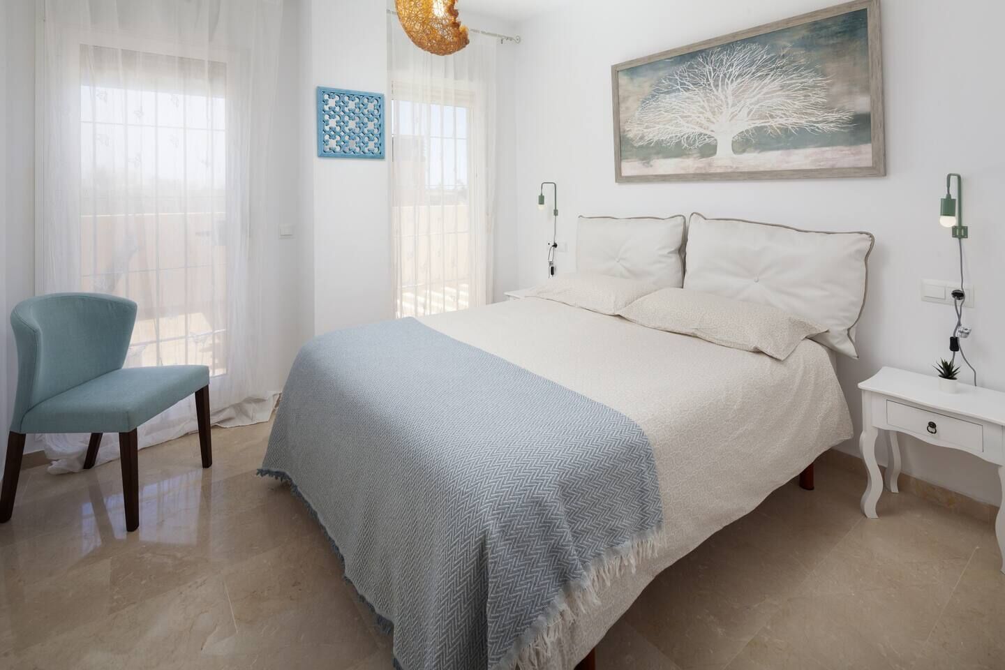 A bedroom with a bed, a chair, a nightstand, a wall art, and a window with sheer curtains.