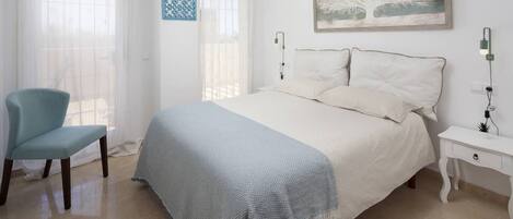 A bedroom with a bed, a chair, a nightstand, a wall art, and a window with sheer curtains.