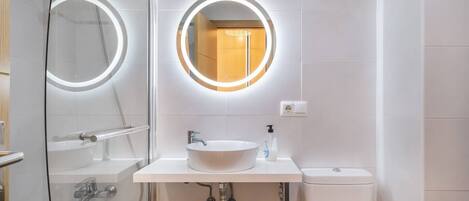 Un baño moderno con ducha de vidrio, espejo redondo con iluminación integrada, lavabo blanco y retrete.