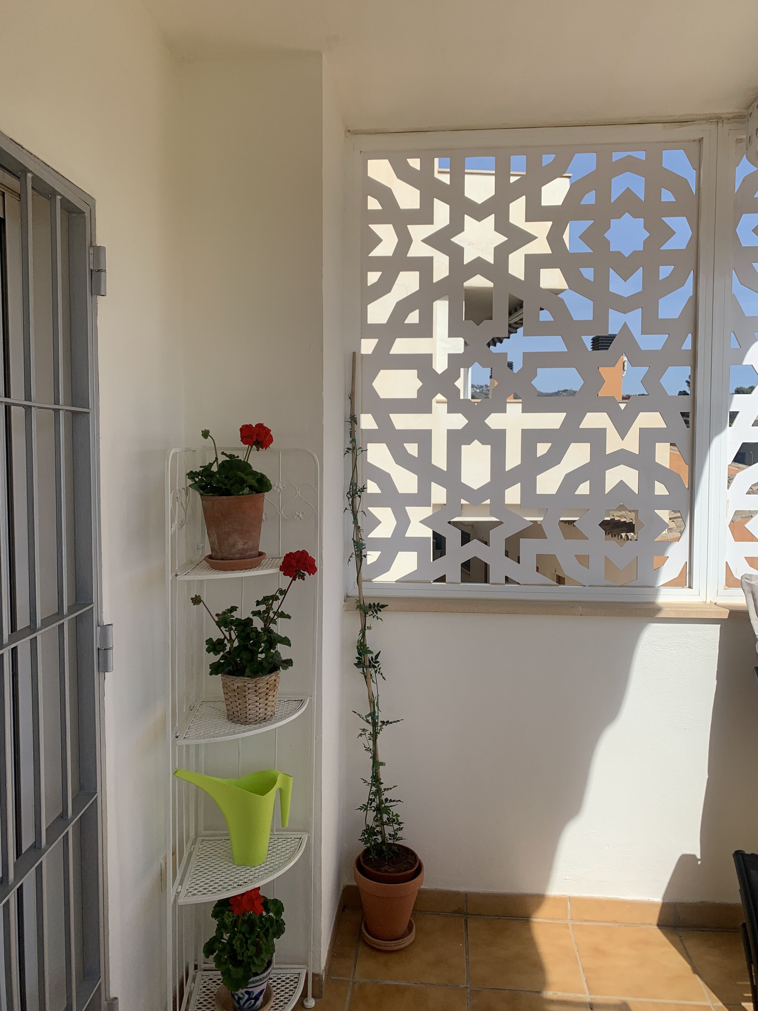 Een klein balkon met een witte metalen plank waarop potplanten staan en een groene gieter. Op de achtergrond is een decoratieve witte scheiding met geometrische patronen zichtbaar.