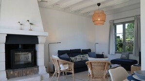 A living room with a fireplace, a sofa, two wicker chairs, a dining table, and a large window with curtains.