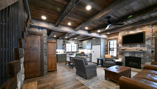 A rustic interior with exposed wooden beams, a stone fireplace, a flat-screen TV mounted on the wall, and a cozy seating area with a leather couch and a chair.