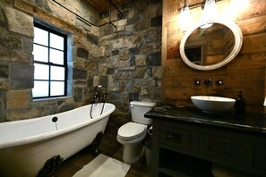 Une salle de bain avec un mur en pierre, une fenêtre, une baignoire, des toilettes et un lavabo avec un miroir.